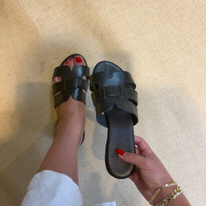 Person wearing black triple-strap sandals, standing on a beige carpet; someone holds the other sandal nearby, showing red nail polish on toes.