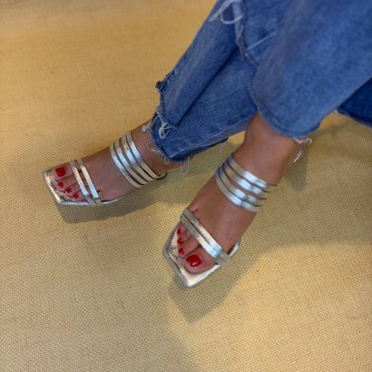 Feet in silver multi-strap sandals with red toenail polish, wearing frayed-hem blue jeans on a beige carpet.