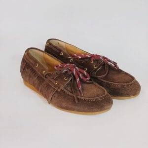Brown suede moccasins with red and white shoelaces resting on a light background, showing a casual, boat-shoe style pair with tan soles
