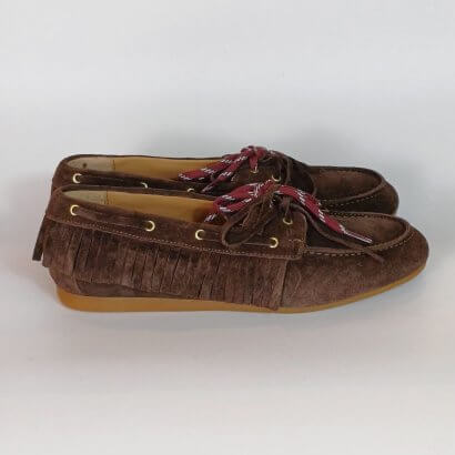 Brown suede moccasin-style loafers with fringe trim and red-and-white laces on a tan sole.