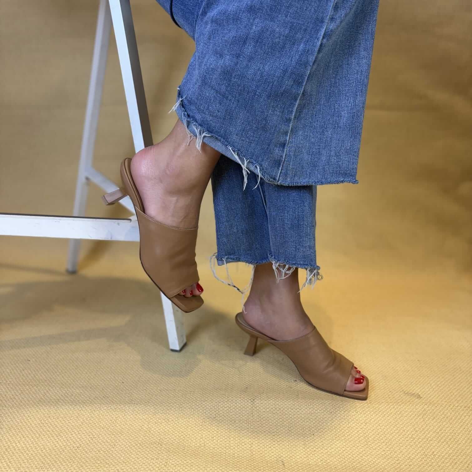 Lower leg and foot wearing frayed-hem blue jeans and tan heeled sandals on a beige carpet, next to a metal step ladder; red toenail polish.